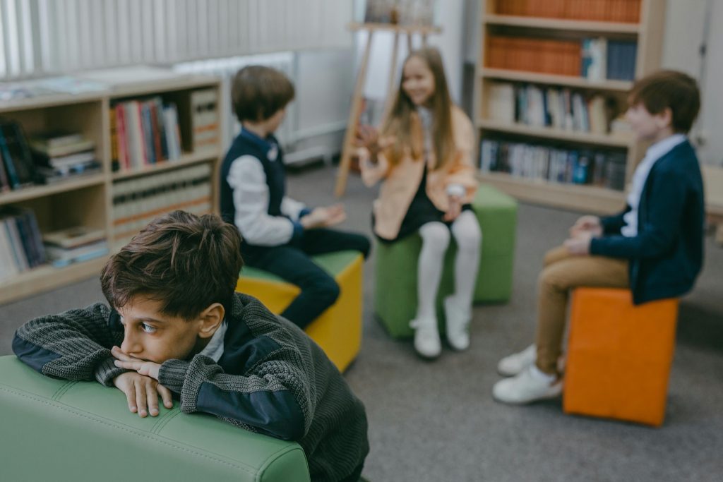 A boy sitting apart while peers talk in the background, reflecting the challenges children with autism build friendships can face in social environments