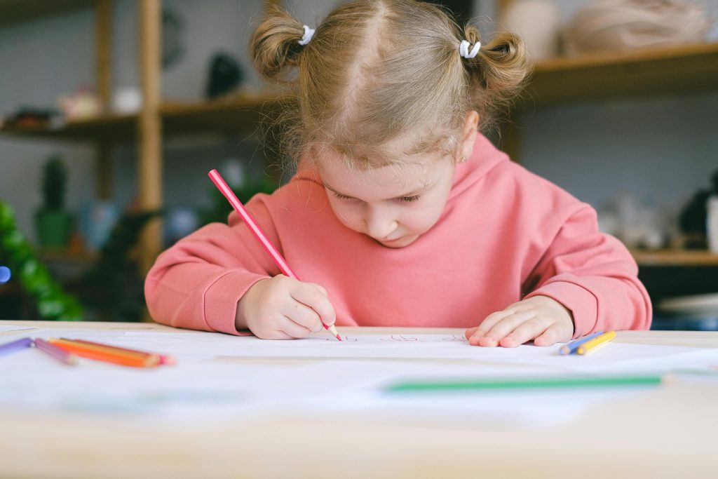 A young girl focusing on writing with colored pencils, showing early learning and how children with autism build friendships through shared classroom activities